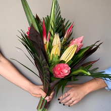Two people's hands holding a Green, pink & yellow tropical bouquet with (2) Ginger Pink, (1) Ginger Red, (2) Chili Pink Picks, (2) Anglonema, (2) Arrow Palm, (6) Cordelyne, (1) Croton Tip Fountain, (2) Masajeana, (2) Pandanus,. 