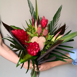2 people holding pink, green, and beige tropical bouquet of (3) Ginger Pink, (2) Sassy, (2) Ananas, (3) Pandanus, (1) Croton Pecoso, (2) Arrow Palm, (3) Xantal, (5) Cordelines Leaves, (2) Masajeanas, (1) Anglonema, (2) Lettuce Ferns.