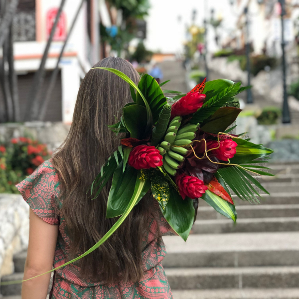 Woman holding a red & green tropical bouquet of (1) Banana Fingers, (4) Ginger Red, (6) Cordelyn, (2) Croto Largo, (3) Palm, (2) Pandanus, (1) Pandanus Curly, (1) Tip Cordelyne babydoll, (3) Willow. 